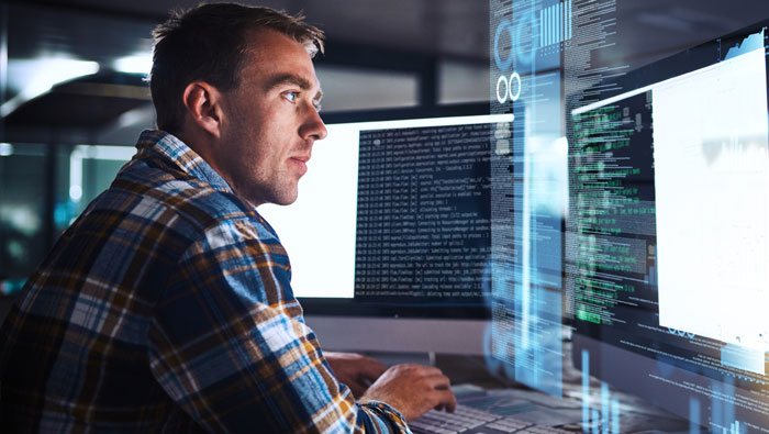 A focused man in a plaid shirt works on a computer, surrounded by multiple screens displaying code and data.
