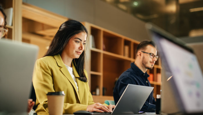 A woman in a yellow blazer works on a laptop, while a man in a dark shirt sits nearby, both in a modern workspace.